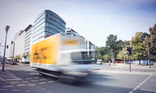 At a junction passes a yellow truck at high speed (motion blurred) with Office buildings in the background and trees on the right.
